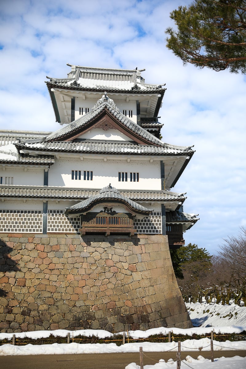 Close-up of Kanazawa Castleโs white plastered tower and massive stone walls overlooking the moat on a sunny winter day.