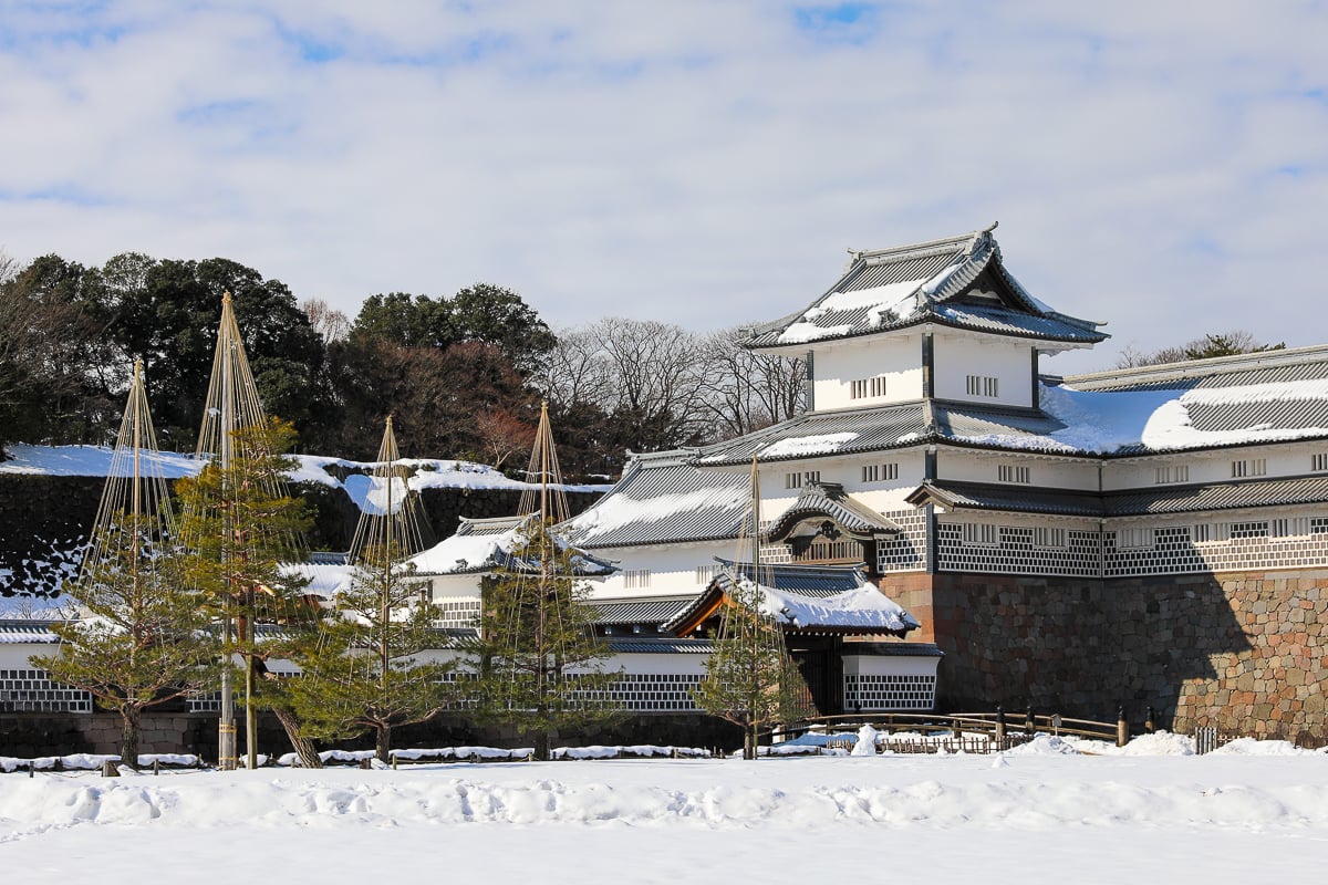 Snow-covered walls and traditional tiled roofs of Kanazawa Castle surrounded by pine trees and winter scenery.