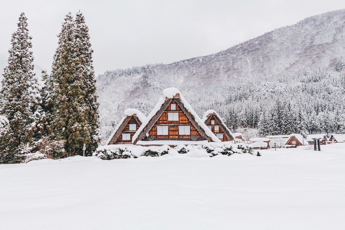Traditional gassho-zukuri farmhouses covered in snow in Shirakawa-go village, Japan, surrounded by snowy mountains and trees.