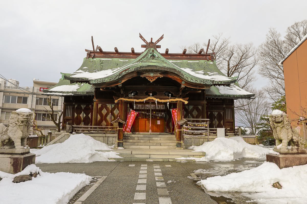 Snow-covered Shinto shrine with stone guardian lions and wooden architecture.