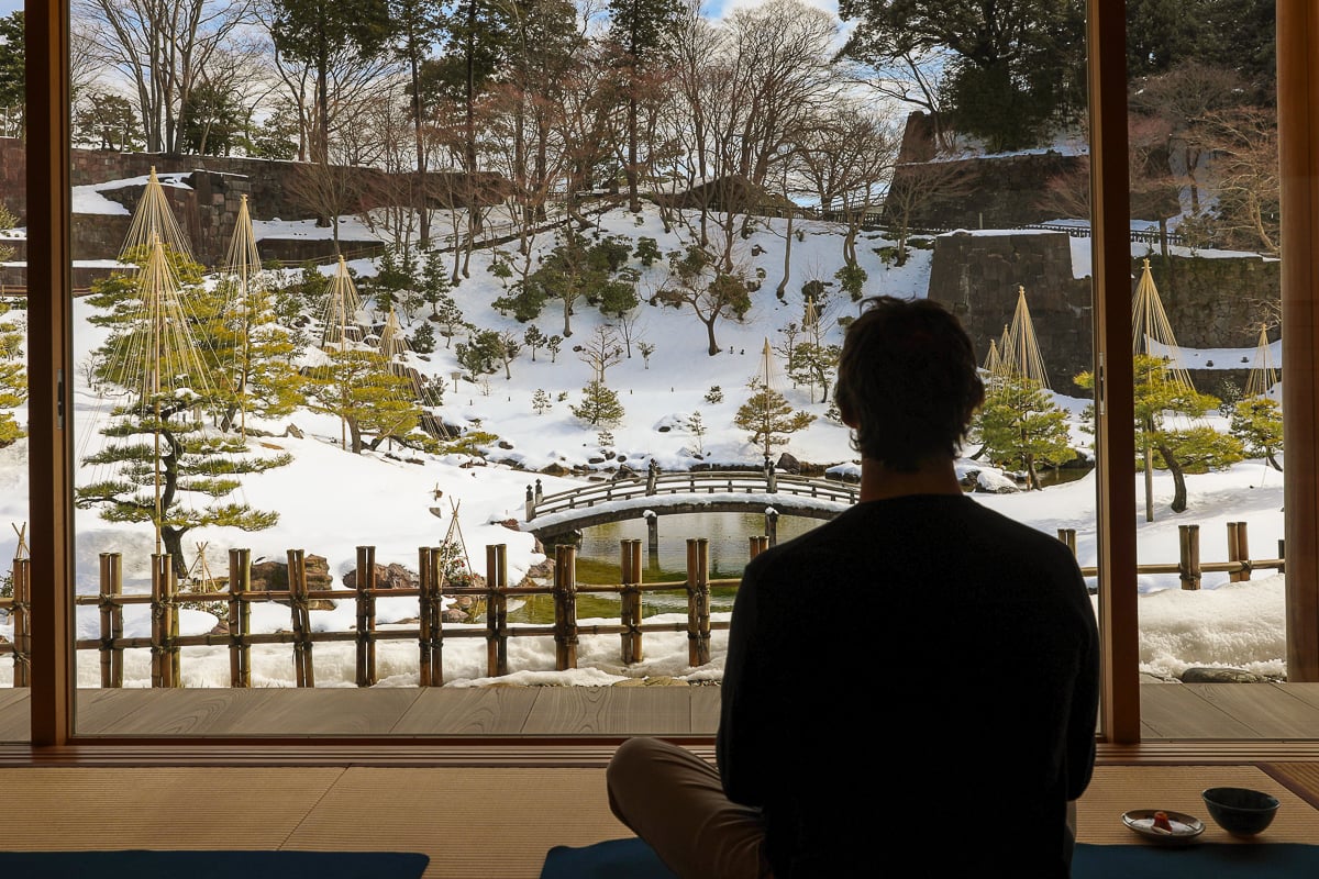 Traveler sitting inside a serene teahouse overlooking a snow-covered Japanese garden and arched wooden bridge in Kanazawa.