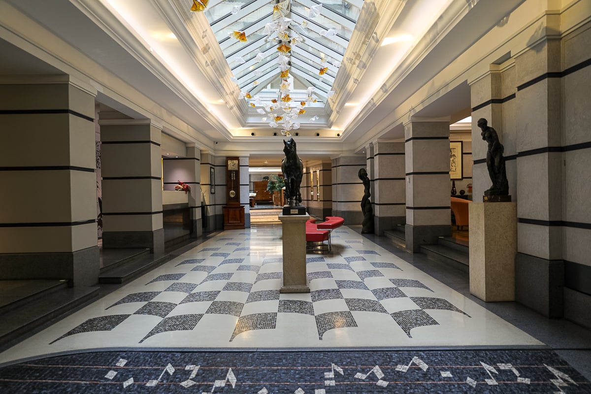 Lobby of the Aria Hotel Prague featuring sculptures, marble floors, and a glass atrium ceiling