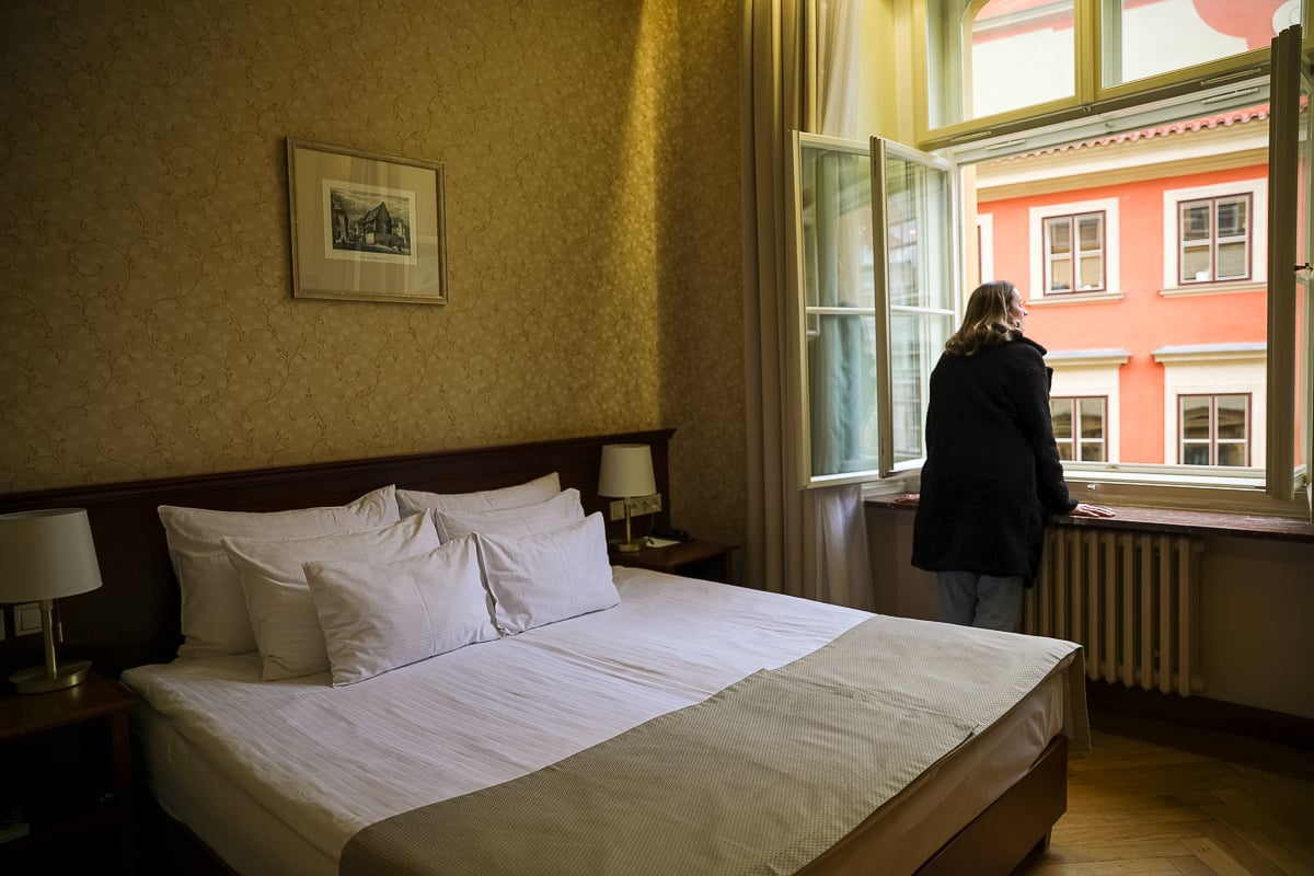 Classic Prague hotel room at the Hotel Rott with a double bed, patterned wallpaper. Maddy is looking through an open window overlooking pastel-colored Old Town buildings.