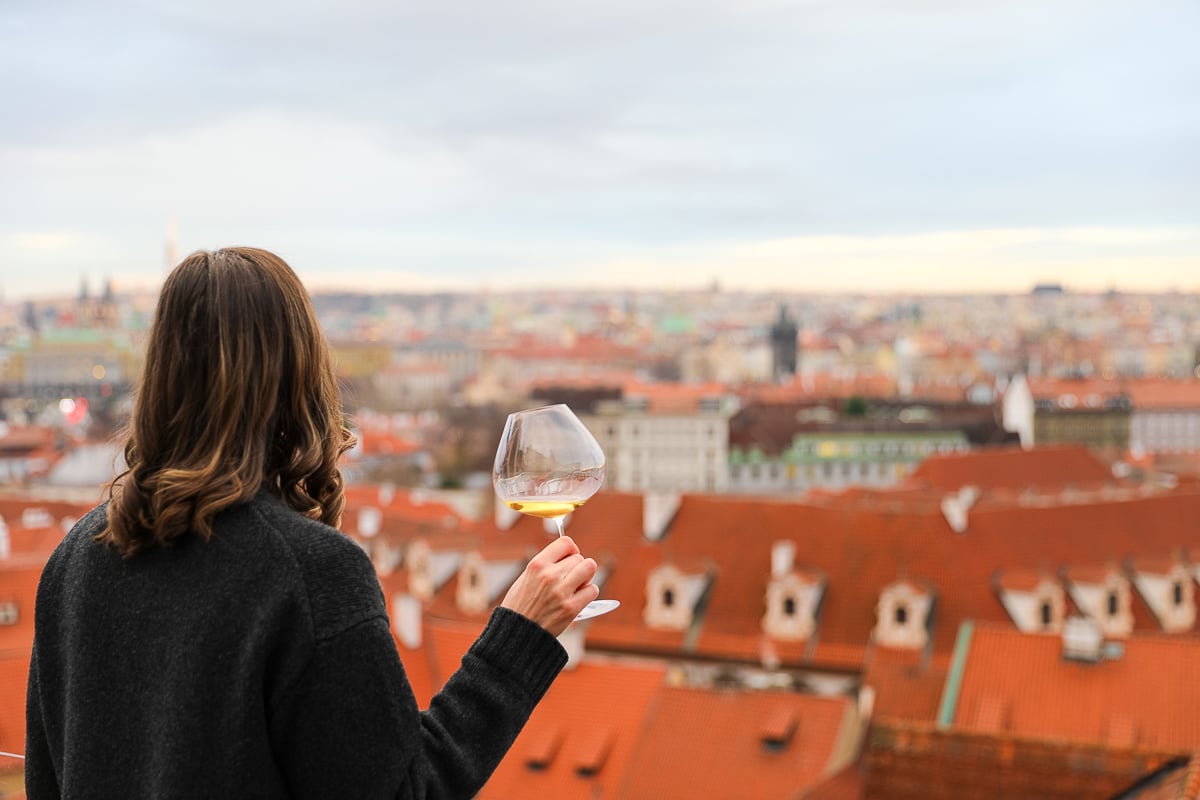 Maddy holding a glass of white wine on the rooftop terrace of Golden Well Hotel, looking out over Prague’s red rooftops and skyline at golden hour