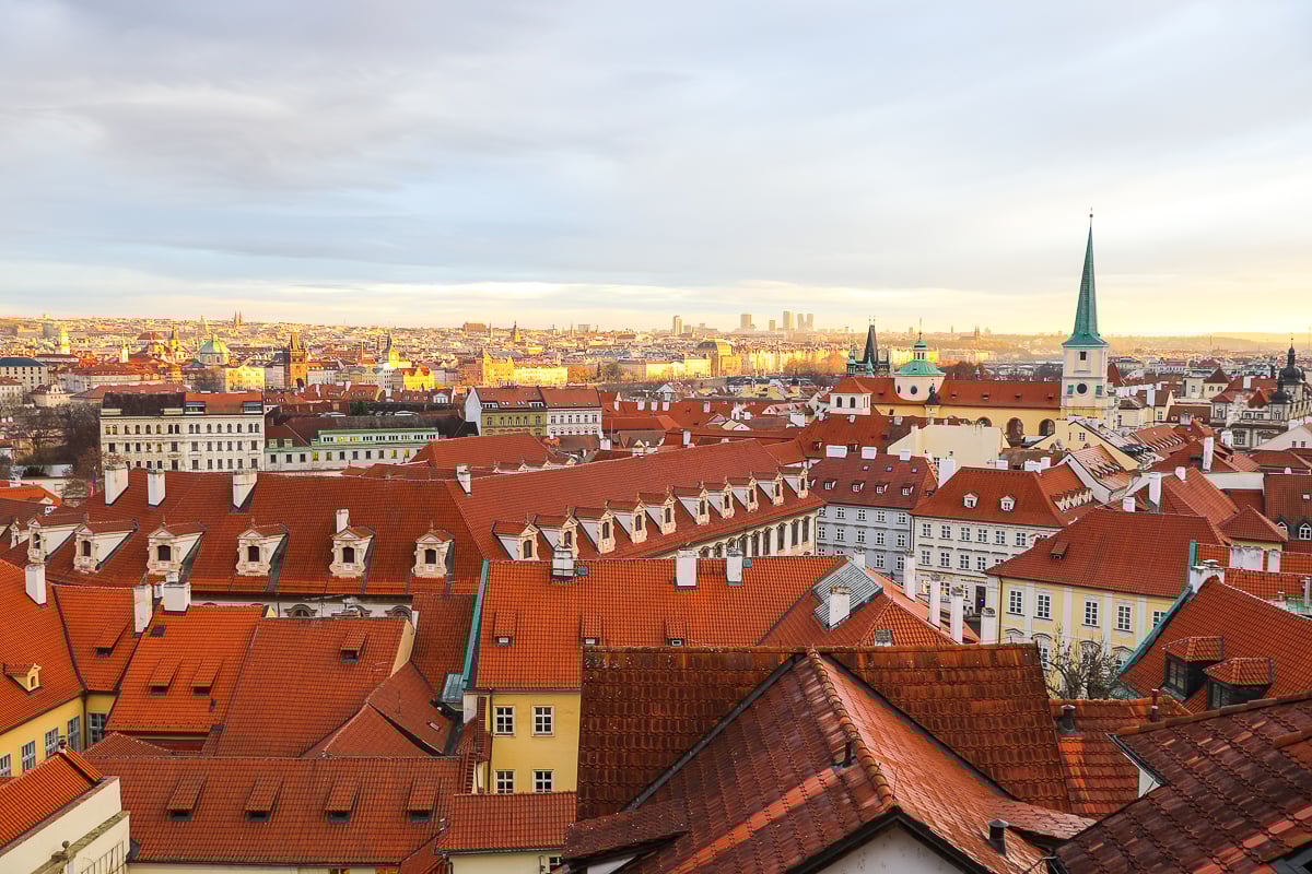 Panoramic rooftop view of Prague’s red rooftops and church spires at sunset, showcasing the cityscape from Malá Strana.