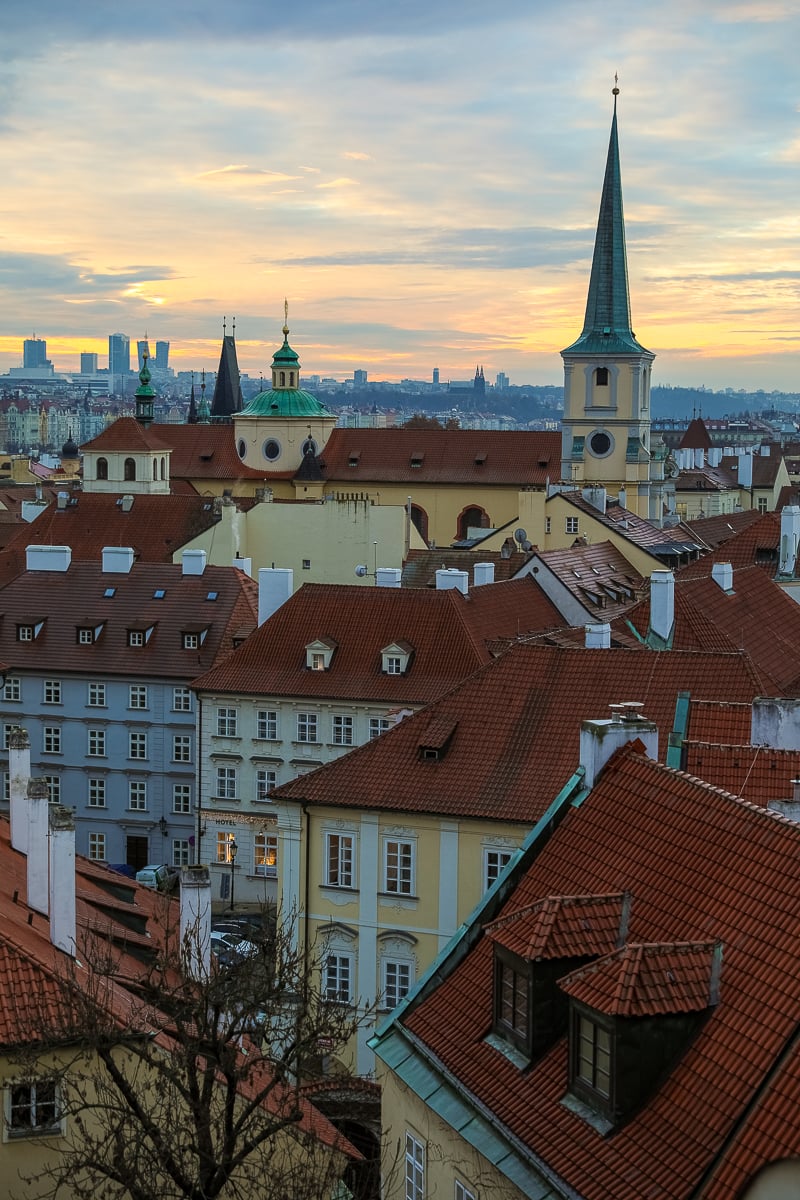 Sunrise view from the Golden Well across Prague rooftops with church towers and pastel buildings.