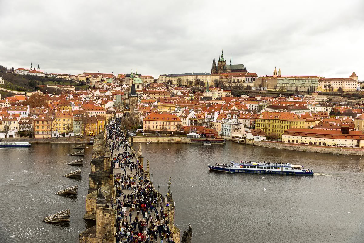 View of Charles Bridge spanning the Vltava River with Prague’s historic skyline, towers, and riverside buildings on a cloudy day