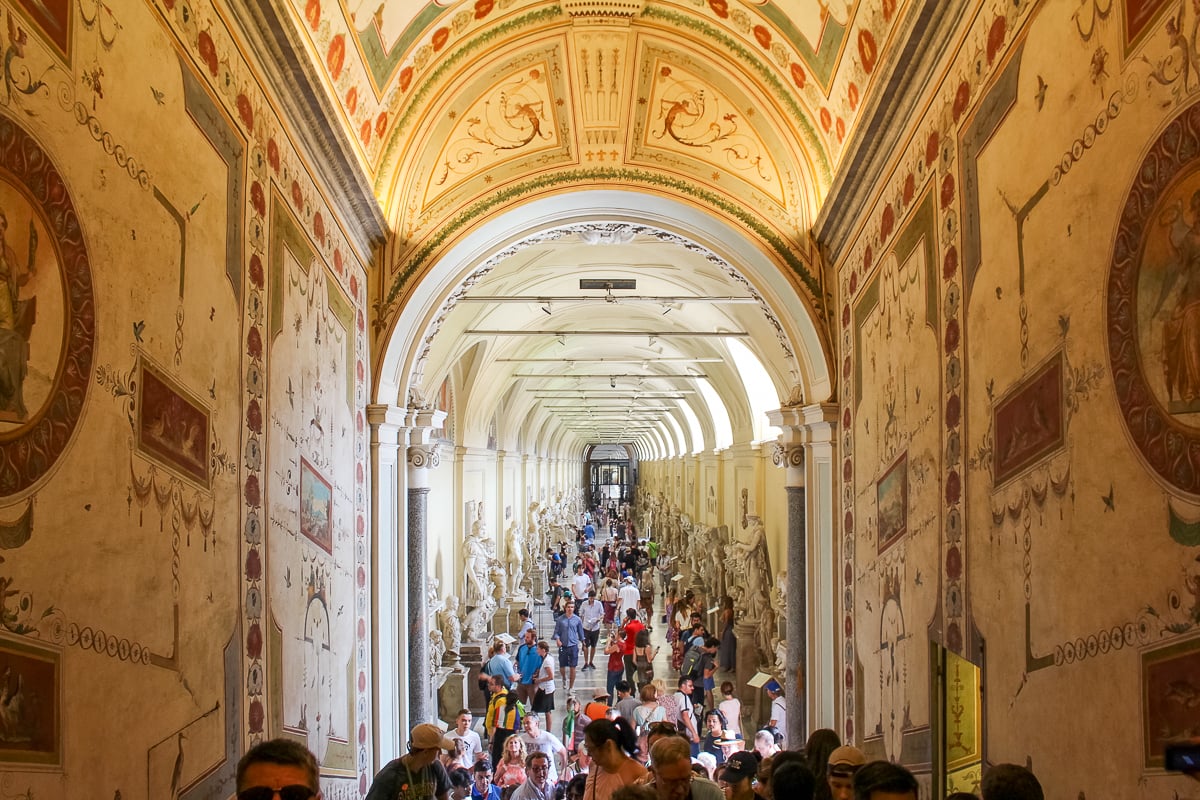 Long arched hallway inside the Vatican Museums lined with marble statues and dense visitor crowds moving through