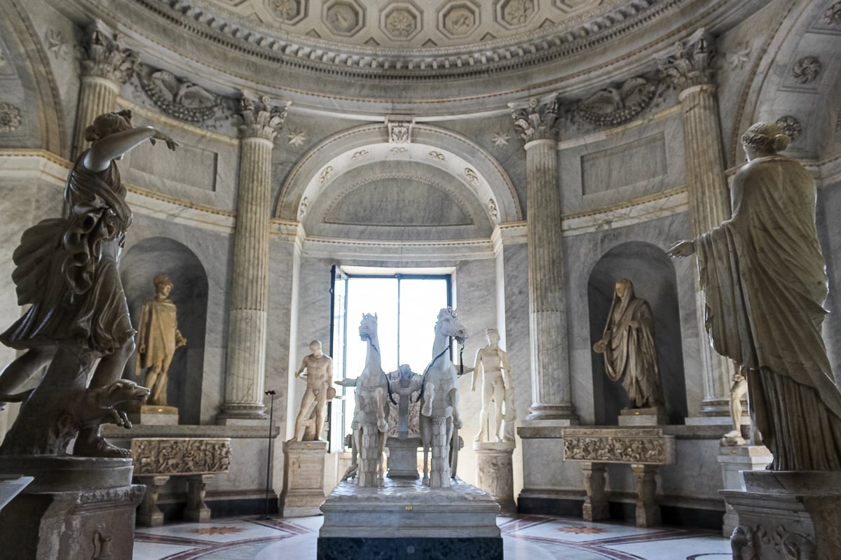 Octagonal Courtyard inside the Vatican Museums with a domed marble room, classical statues in wall niches, and a central sculpture of two horses framed by tall columns and natural light