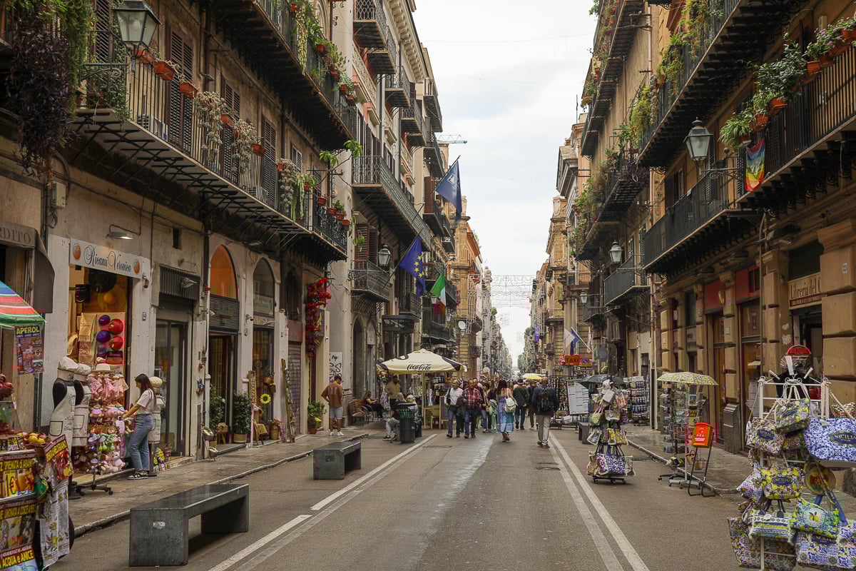 Lively Palermo walking street with historic buildings, balconies, shops, and crowds in Sicily