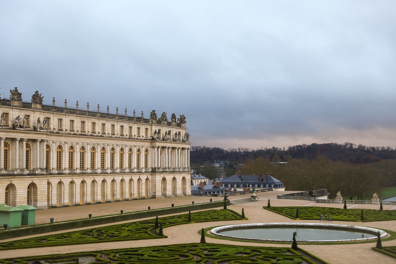 View of Versailles Palace gardens during a day trip from Paris with manicured hedges and reflecting pool under a cloudy sky