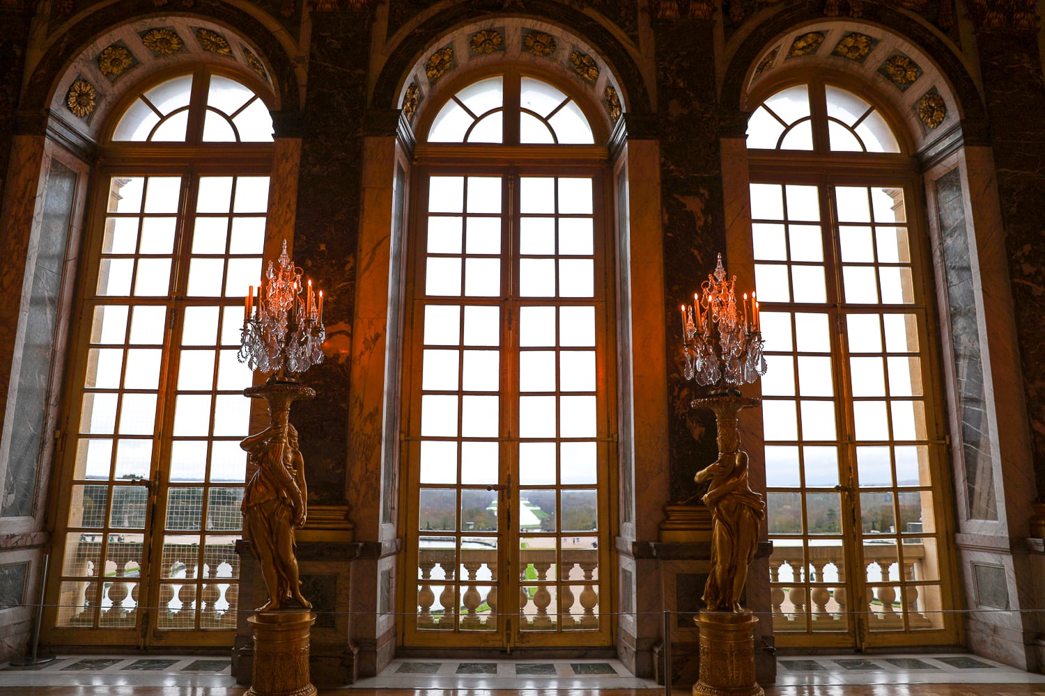 Tall arched windows inside Versailles Palace with marble walls and candlelit crystal lamps