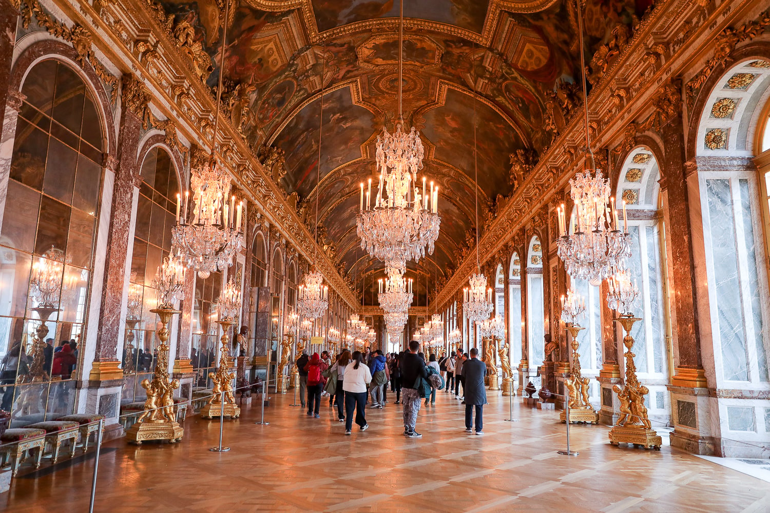 Hall of Mirrors at Versailles Palace with chandeliers, gilded arches, and mirrored walls reflecting the crowd