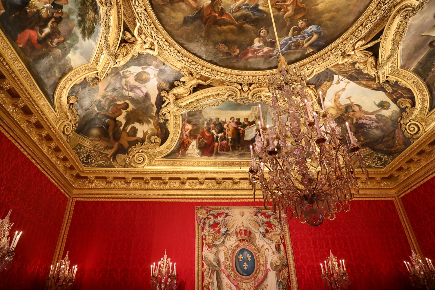 Versailles Palace salon with red damask walls, painted ceiling frescoes, and a crystal chandelier