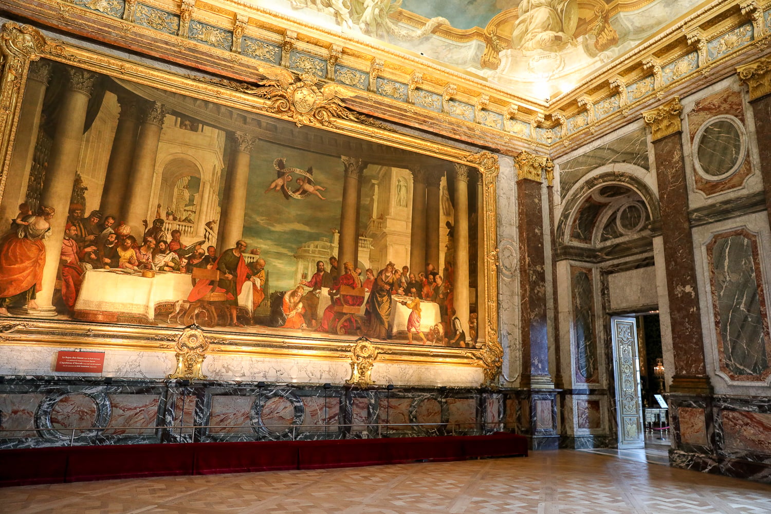 Ornate Versailles Palace gallery with a massive gilded painting, marble walls, and arched doorway