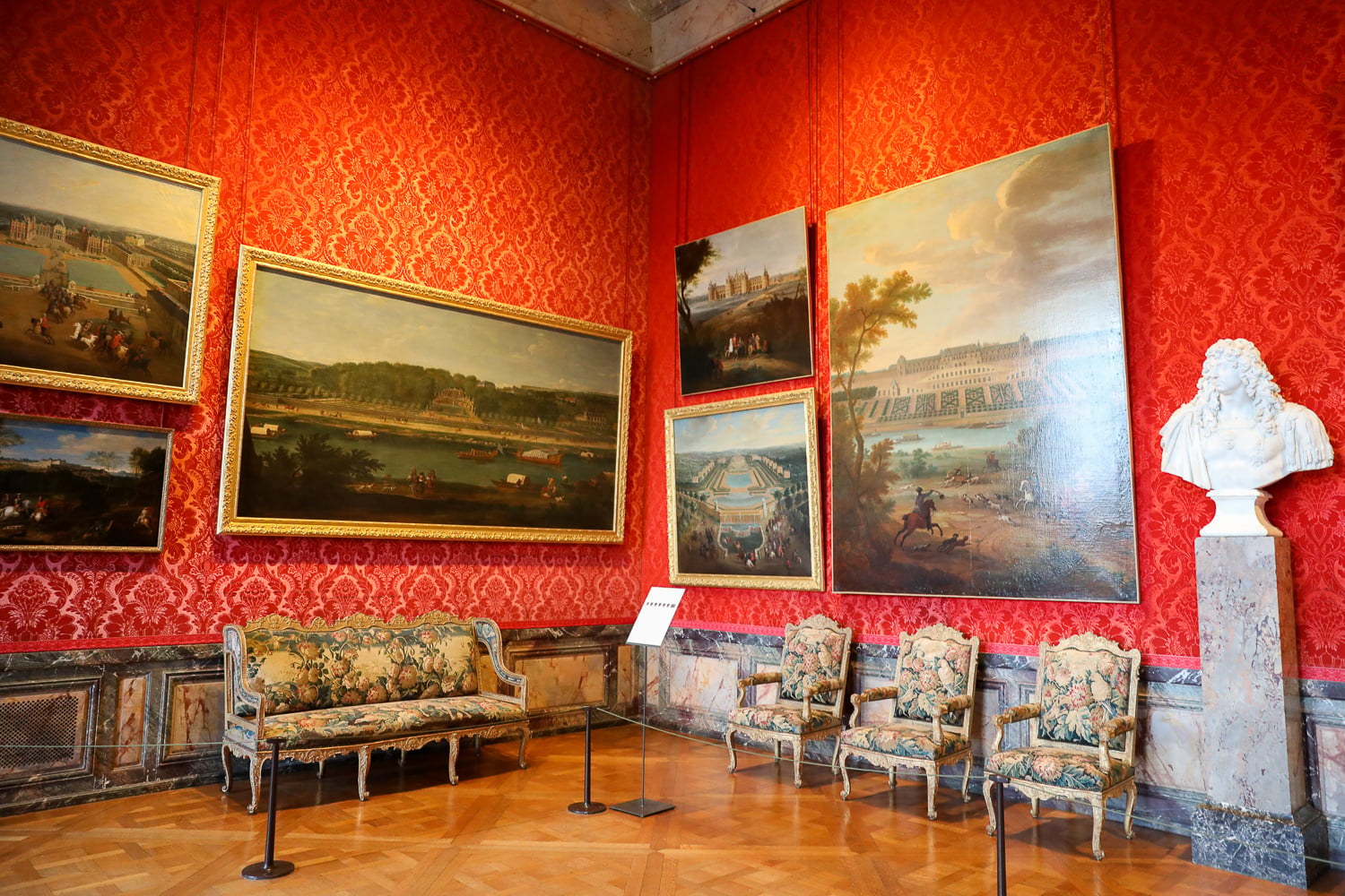 Red damask room in Versailles Palace with framed paintings, antique chairs, and a white marble bust