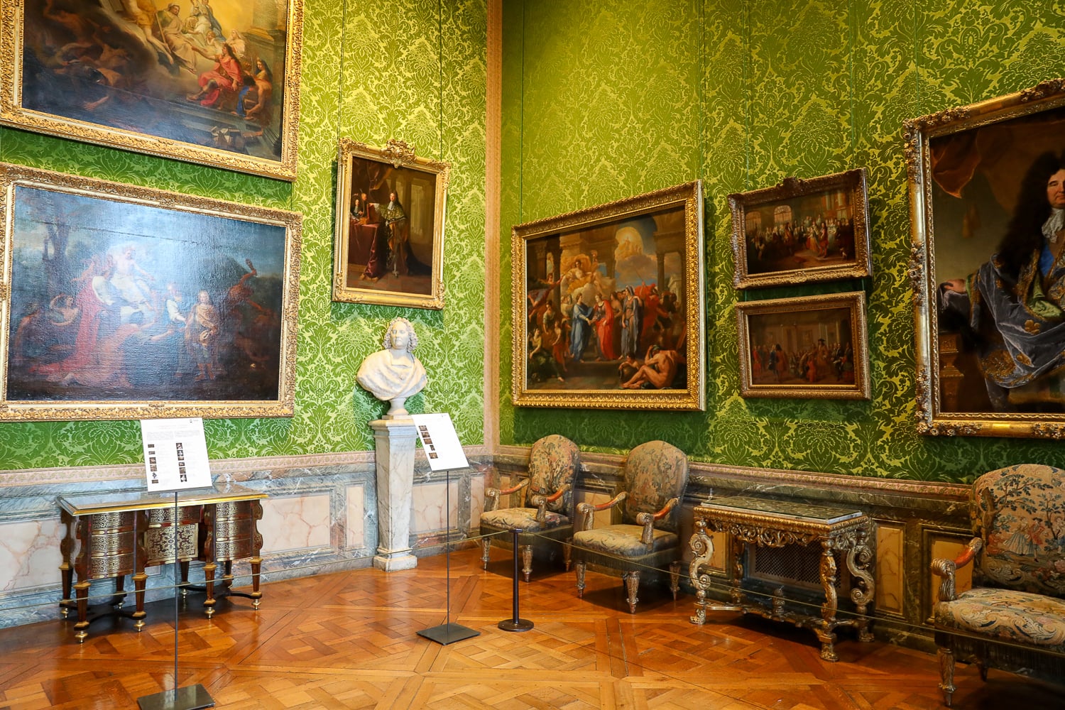 Green damask salon in Versailles Palace with gilded framed paintings, antique chairs, and a marble bust