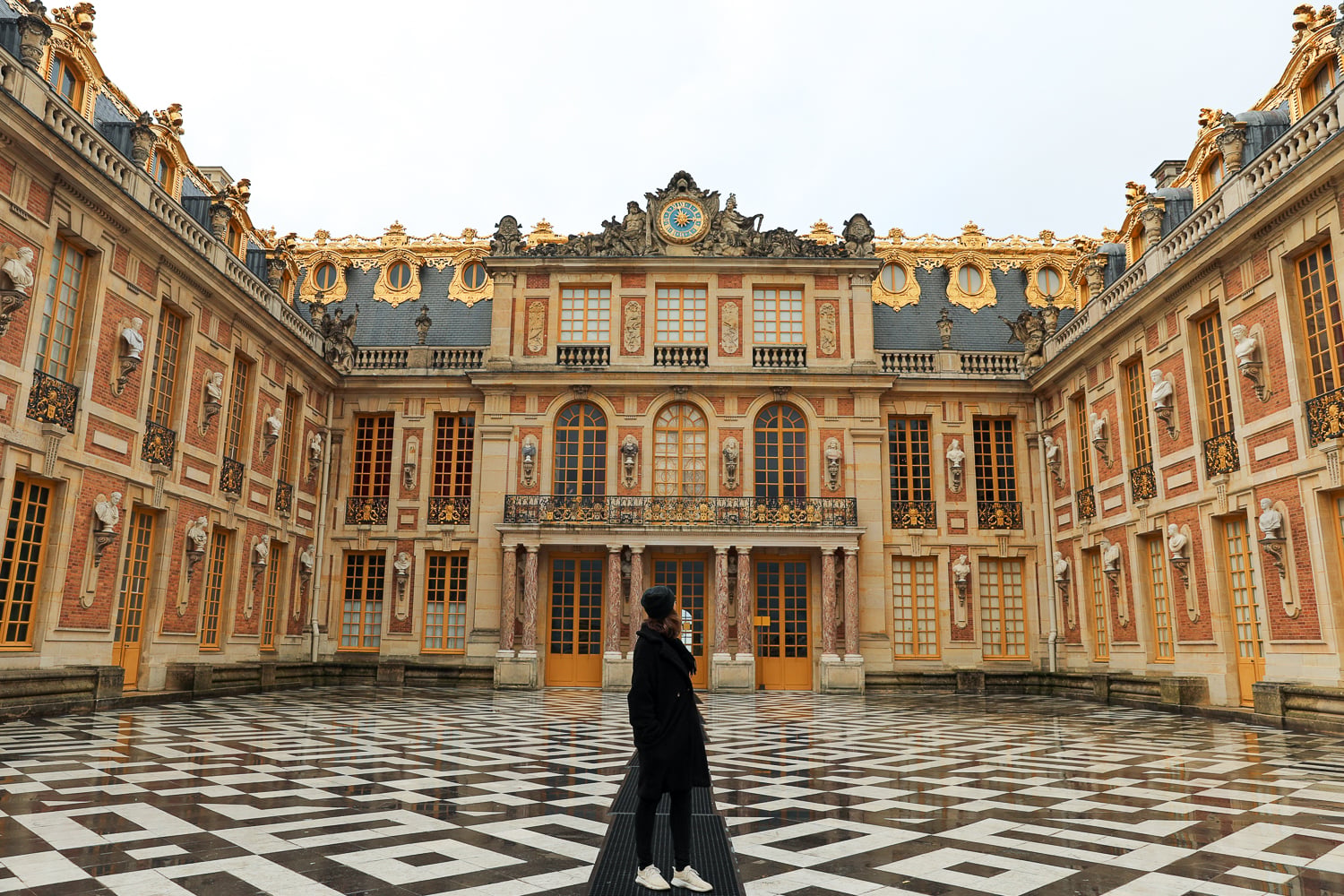 Visitor standing in Versailles Palace’s Marble Courtyard with ornate gold-trimmed façades and a black-and-white stone courtyard