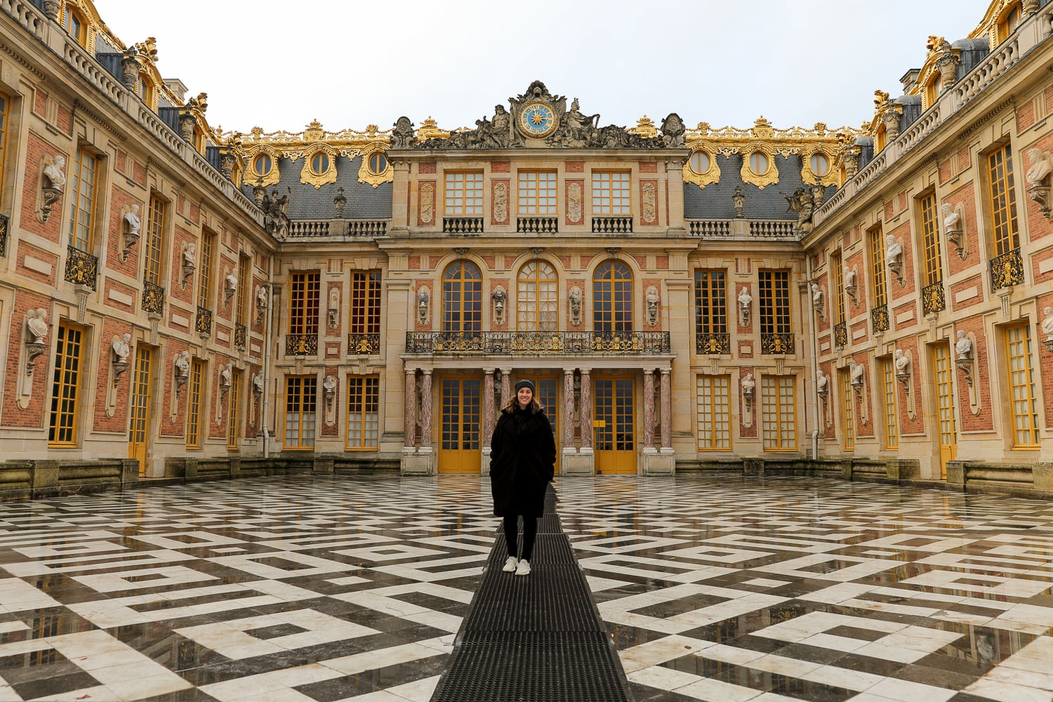 Visitor smiling in the Marble Courtyard at Versailles Palace, framed by gold-trimmed windows and patterned stone flooring