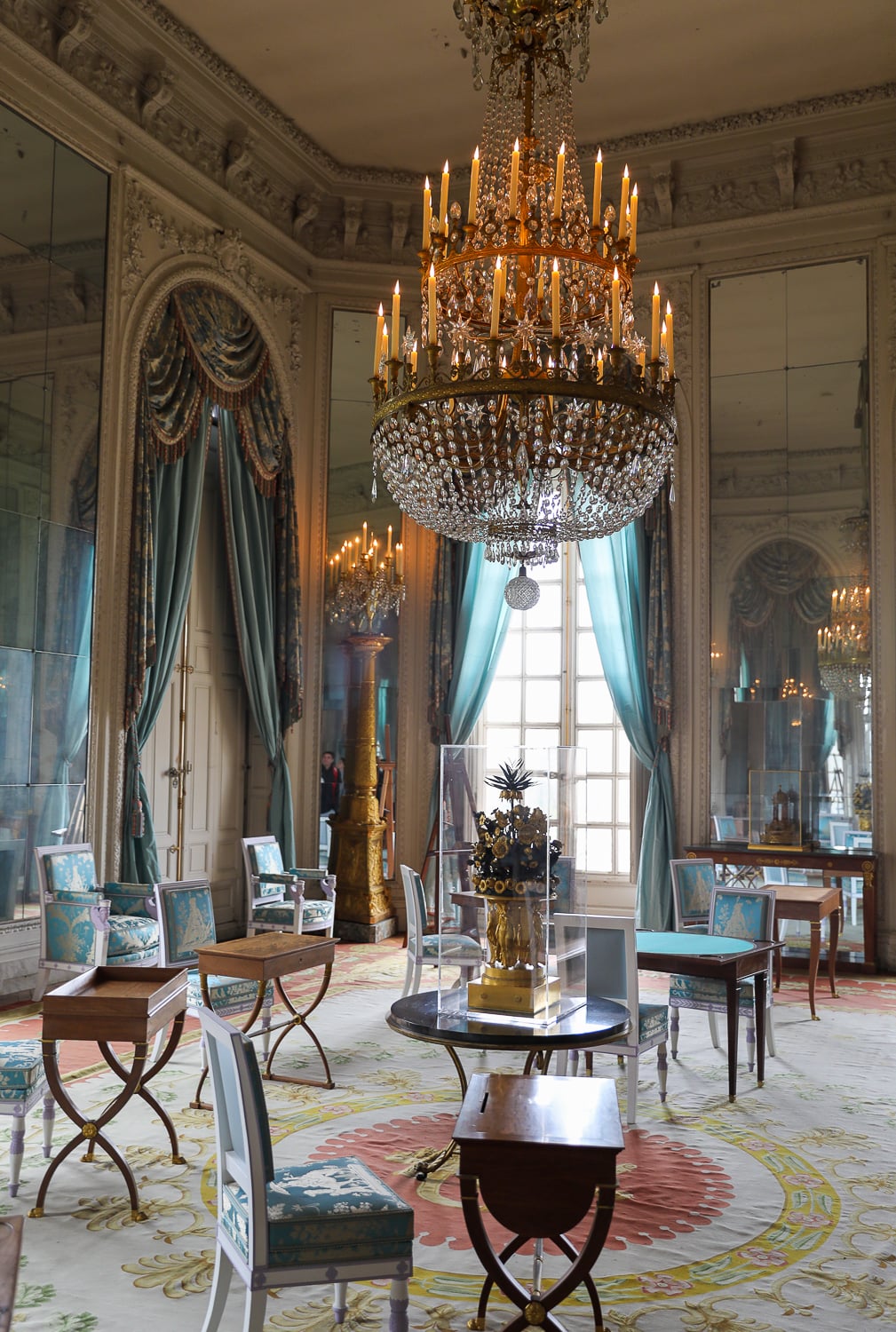 Elegant Versailles Palace salon with a large crystal chandelier, pale blue drapes, and antique chairs and tables
