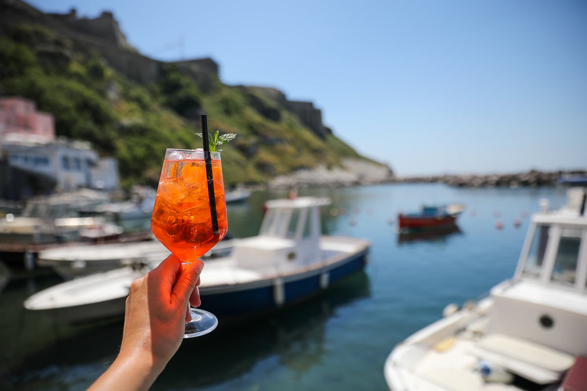 Hand holding an Aperol Spritz over a boat-filled harbor, a classic aperitivo in Procida, Italy