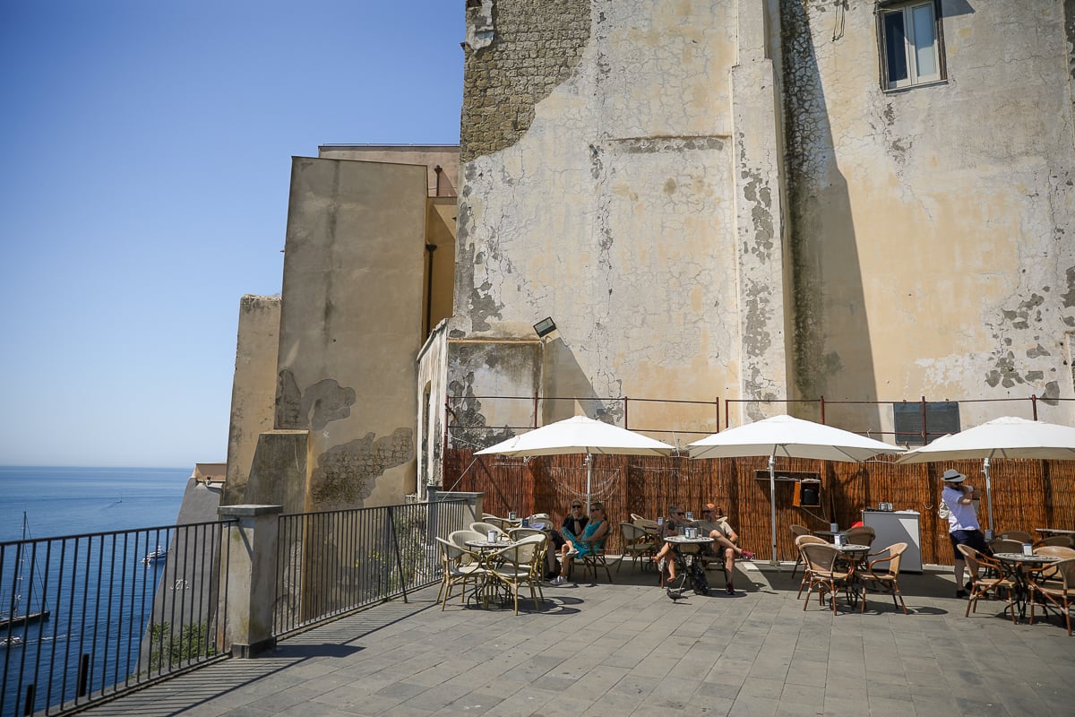 Café terrace with white umbrellas and sea views beside weathered stone walls at Terra Murata, Procida