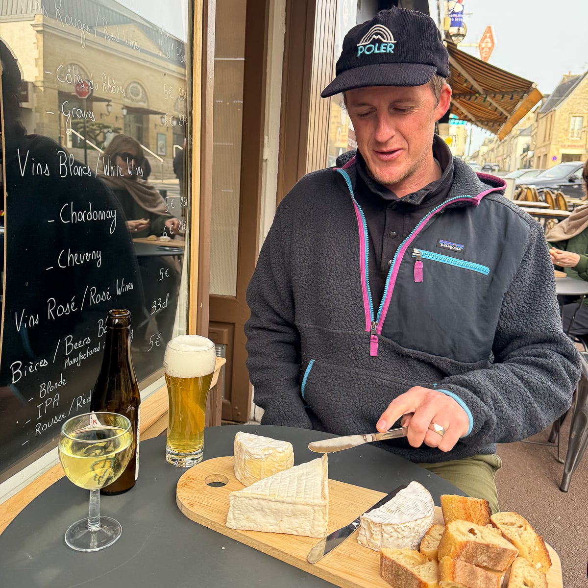Man cutting into a Norman cheese board with local beer and white wine at a pavement café in Bayeux, France