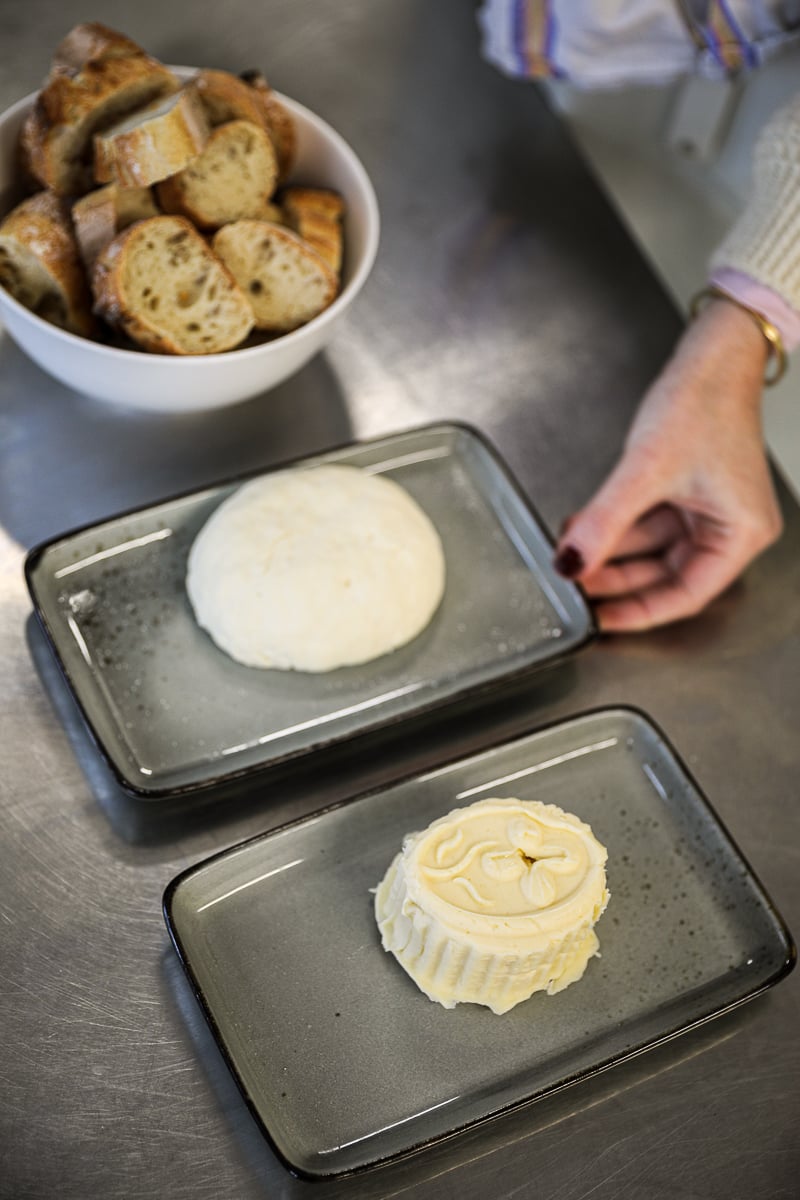 Two rounds of freshly made cheese on slate plates beside a bowl of sliced baguette, one shaped in a flower-stamped mold.