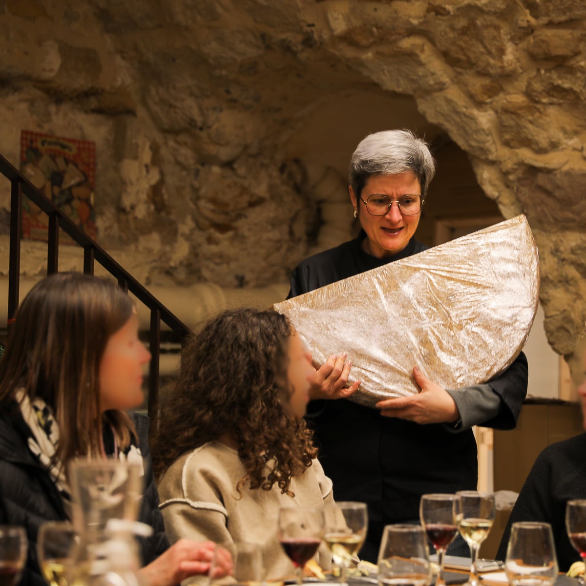 Cheesemonger holding a large wheel of cheese while presenting it to guests seated at a table during a Paris cheese and wine tasting in a stone cellar.