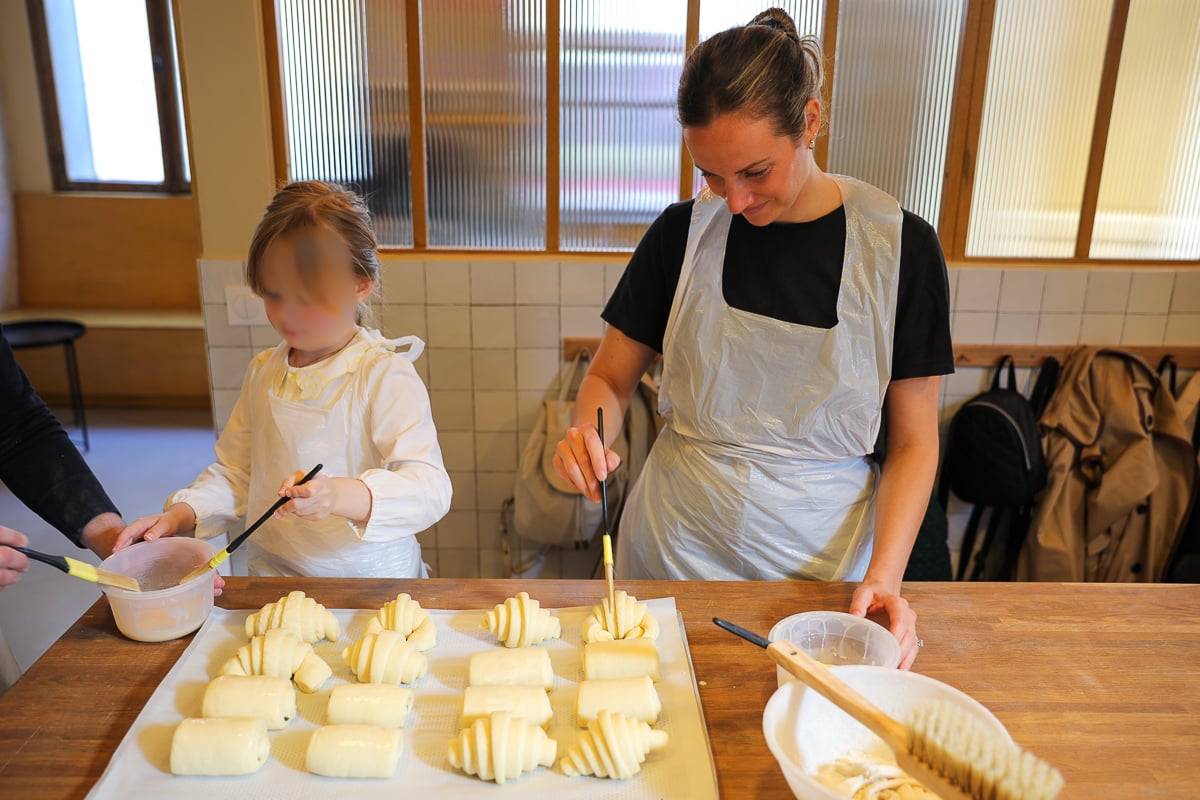 Woman and a young child brushing egg wash onto shaped croissants and pains au chocolat at a family-friendly Paris baking class.