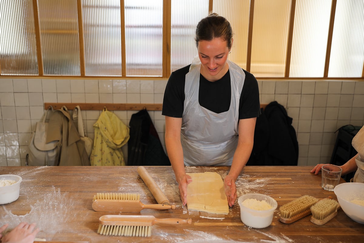Woman smiling as she works laminated croissant dough on a floured table lined with rolling pins and brushes.