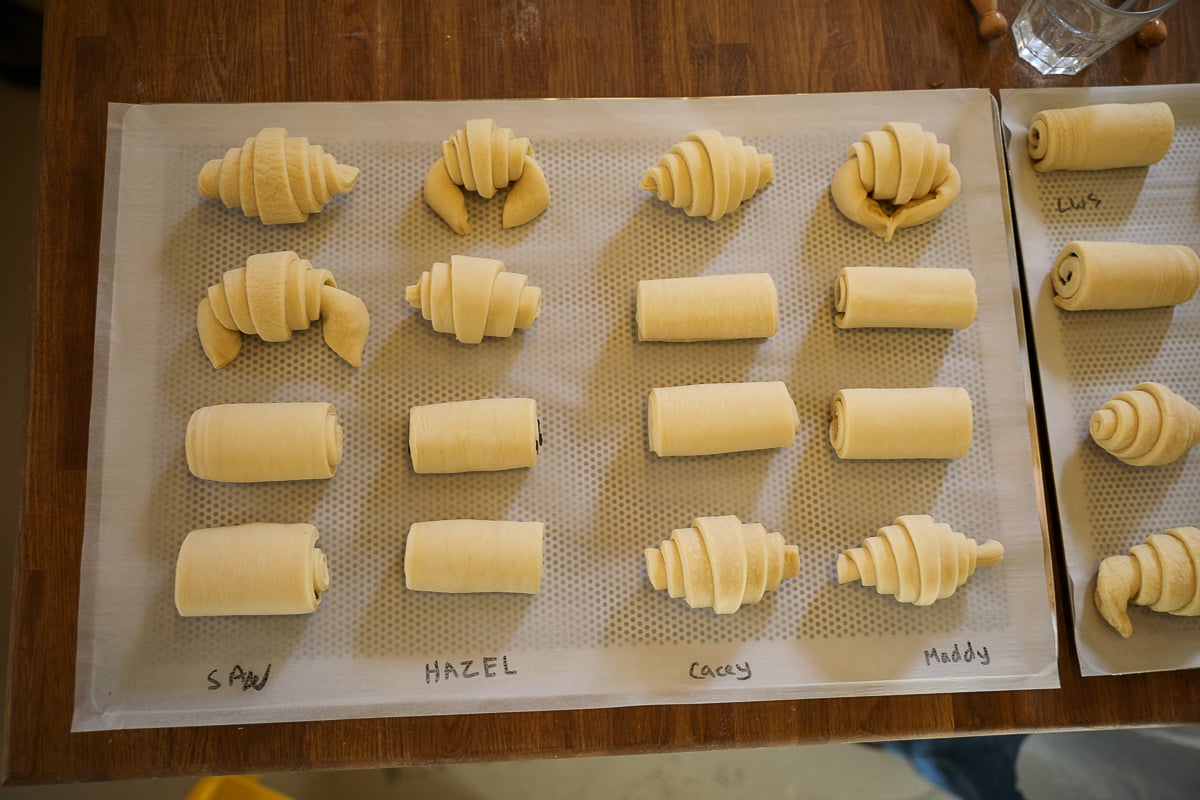 Tray of hand-shaped raw croissants and pains au chocolat labeled with students' names, ready to bake.