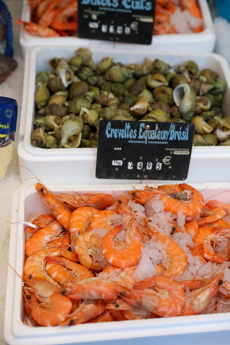 Fresh prawns and sea snails on ice at a Paris market fish stall, with a handwritten price sign.