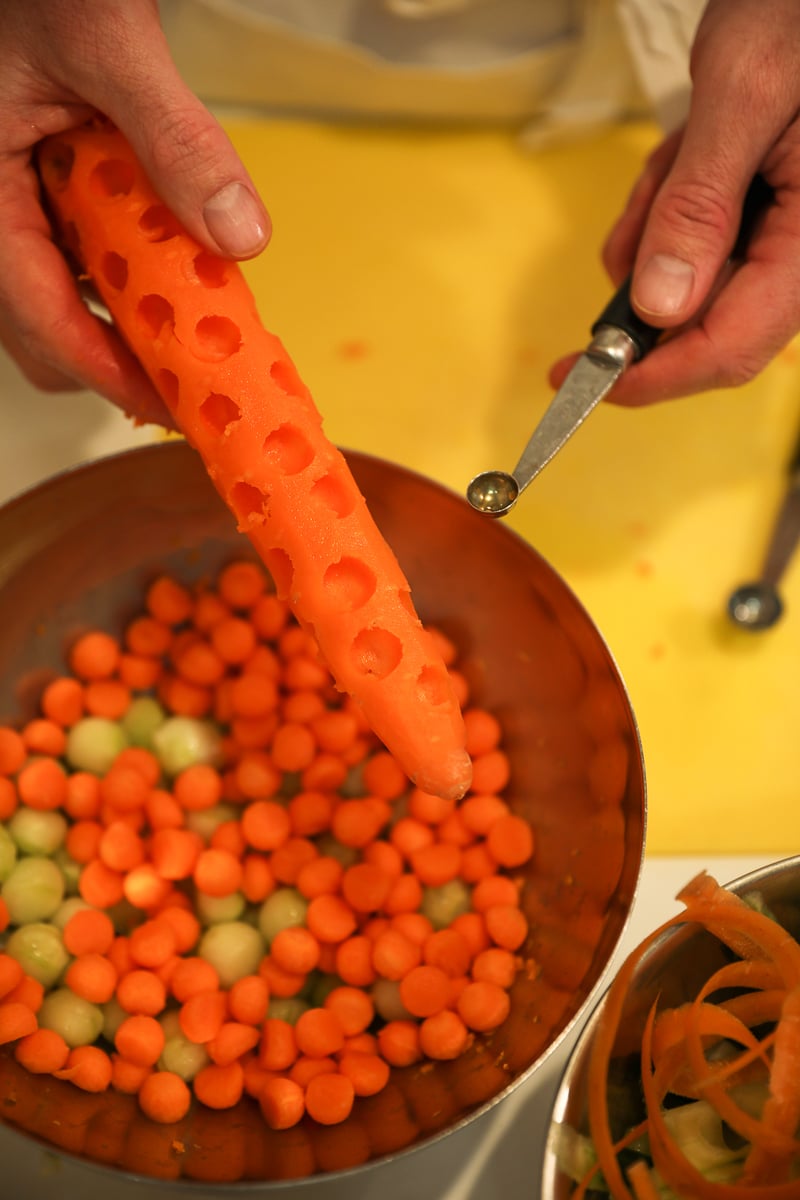 Hands using a Parisienne scoop to carve a carrot into small balls over a bowl of carrot and cucumber spheres.