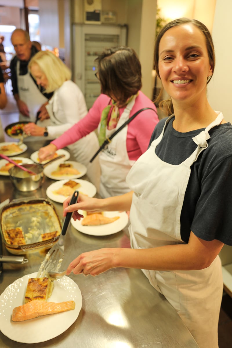 Woman in an apron plating baked salmon and potato gratin alongside other students at a Paris cooking class.