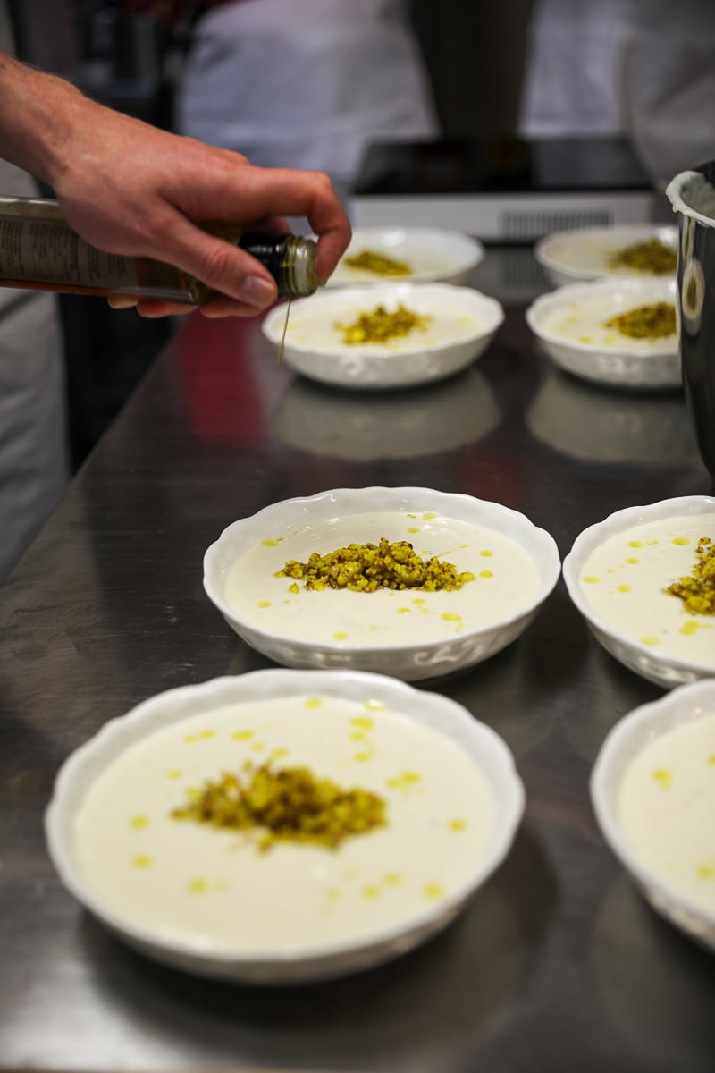 Student drizzling olive oil over bowls of cauliflower velouté lined up on a stainless steel counter.
