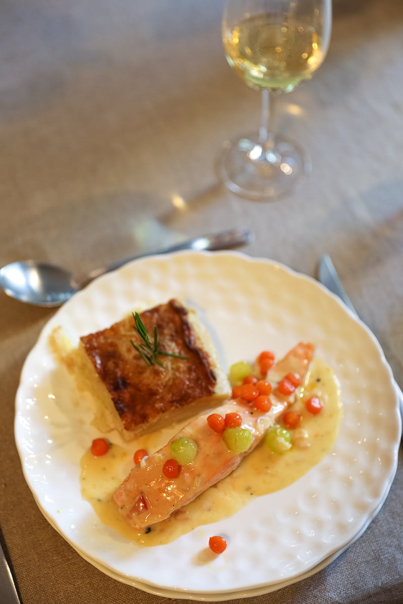 Plated baked salmon with citrus beurre blanc, glazed vegetable balls, and a square of potato gratin, served with white wine.
