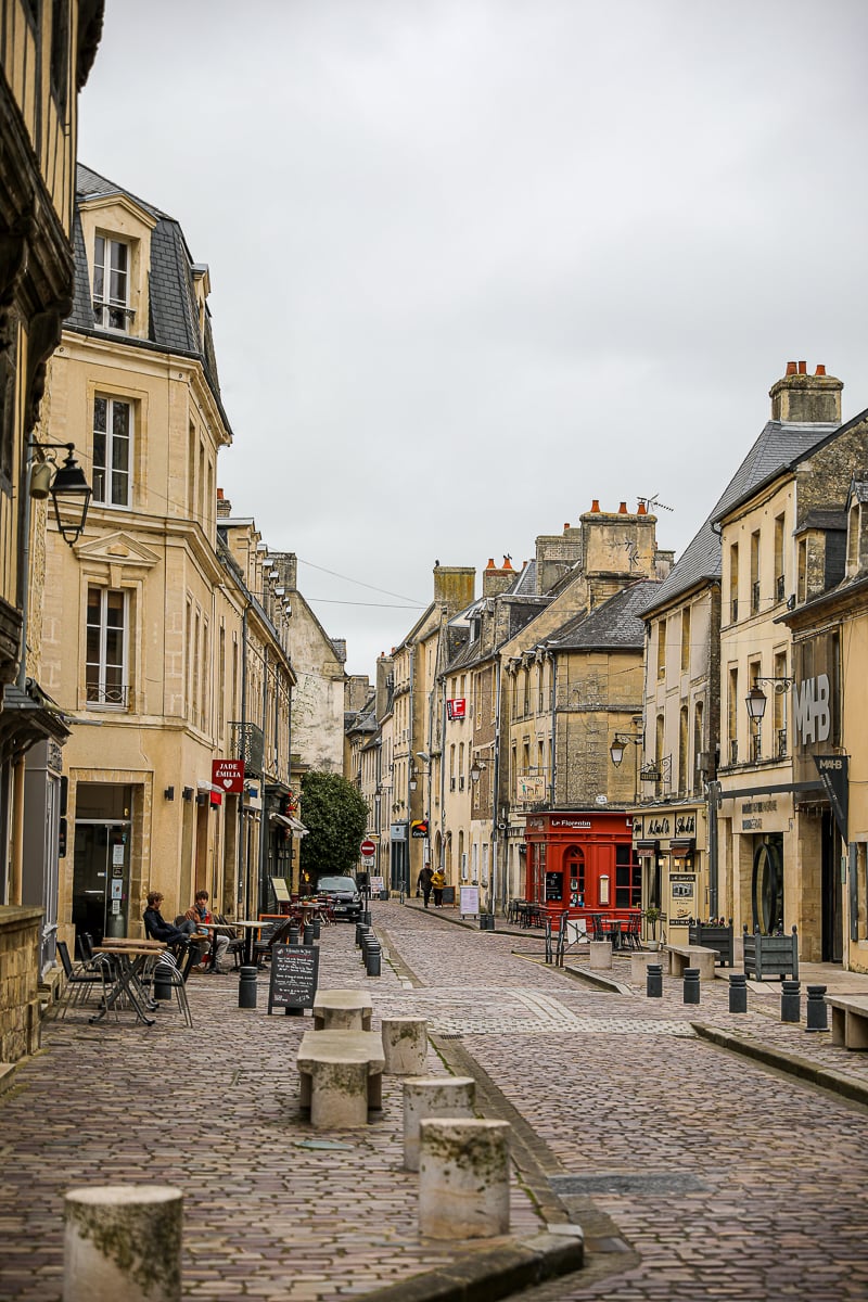 Quiet cobblestone street lined with stone buildings and outdoor café tables in the historic center of Bayeux, Normandy