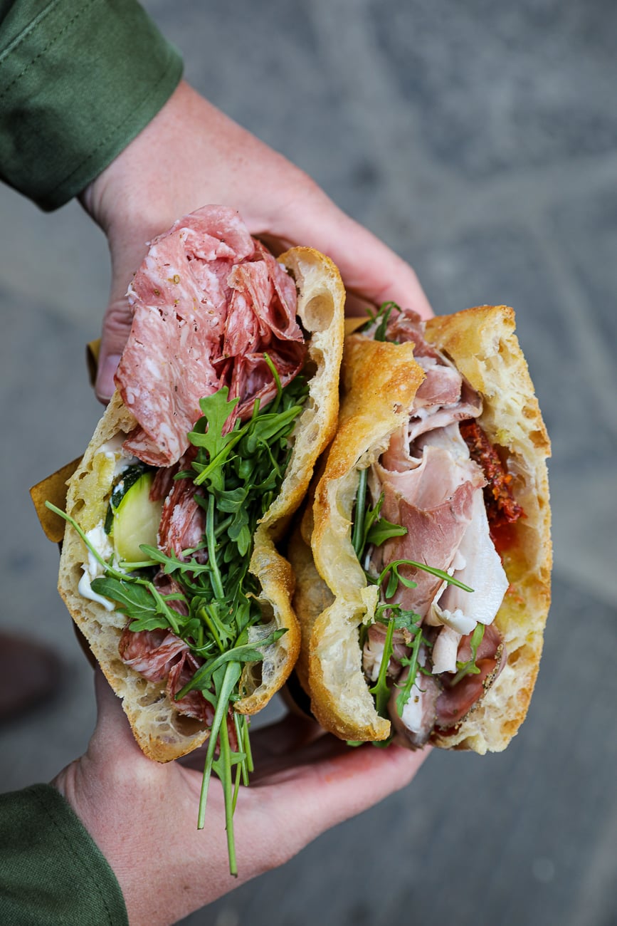 Two schiacciata sandwiches filled with salame, prosciutto, arugula, and sun-dried tomatoes held in hand.