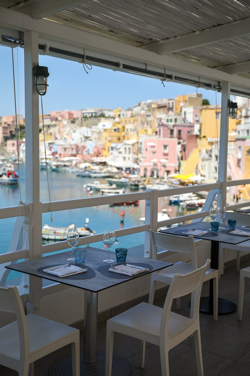 White tables set on a terrace overlooking the colorful Corricella harbor