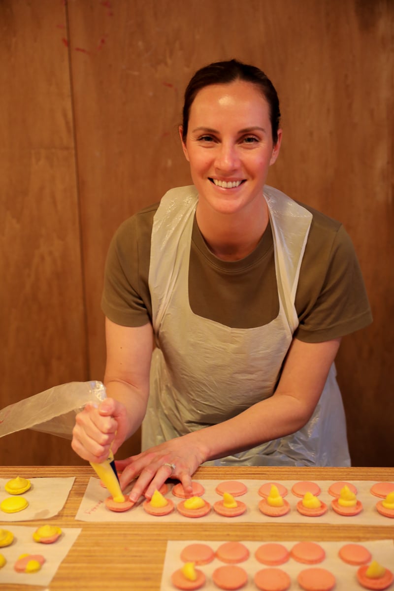 Woman in an apron smiling as she pipes mango filling onto pink macaron shells at a Paris macaron baking class.