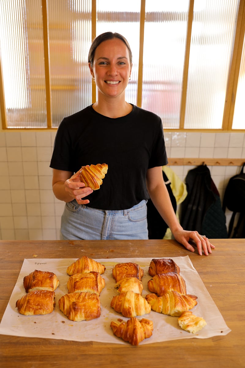 Woman holding up a freshly baked croissant behind a tray of her homemade croissants and pains au chocolat.
