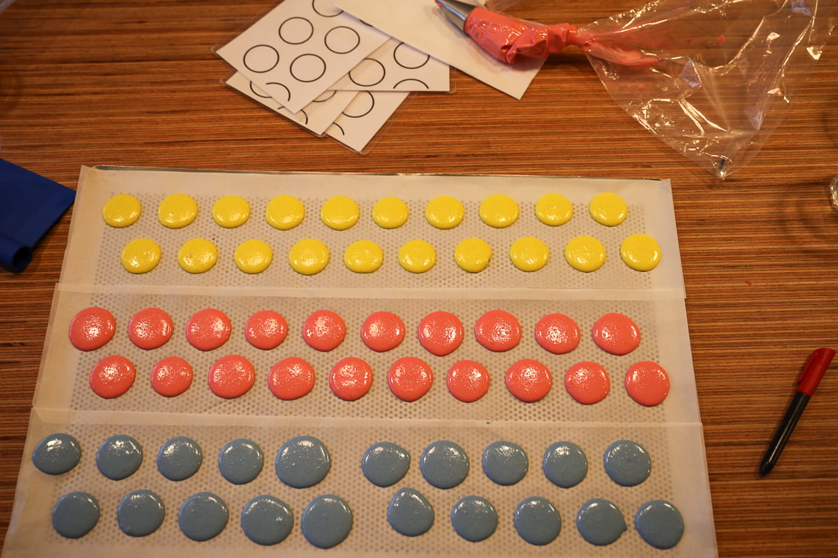 Trays of piped yellow, pink, and blue macaron shells resting on baking paper beside circle piping guides.