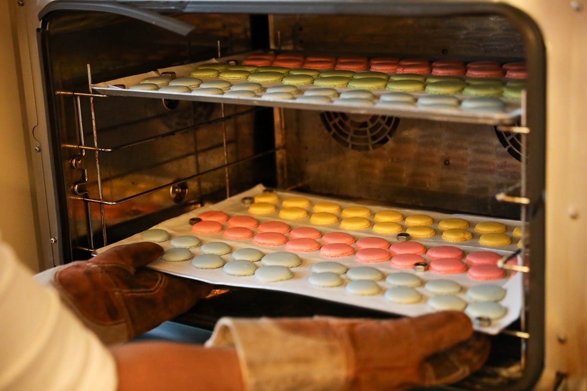 Hands in oven mitts pulling a tray of pastel macarons from the oven during a Paris baking class.