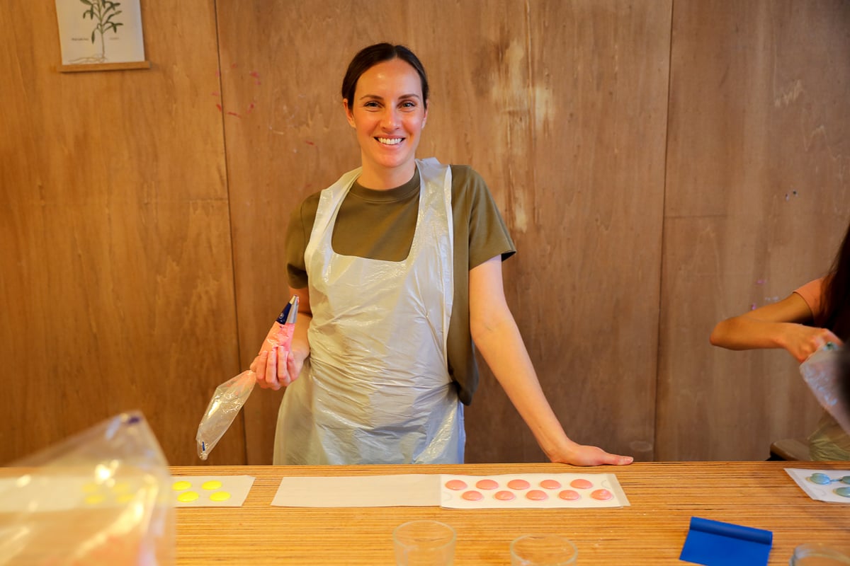Woman in an apron smiling and holding a piping bag at her macaron station, trays of pink and yellow shells in front of her.