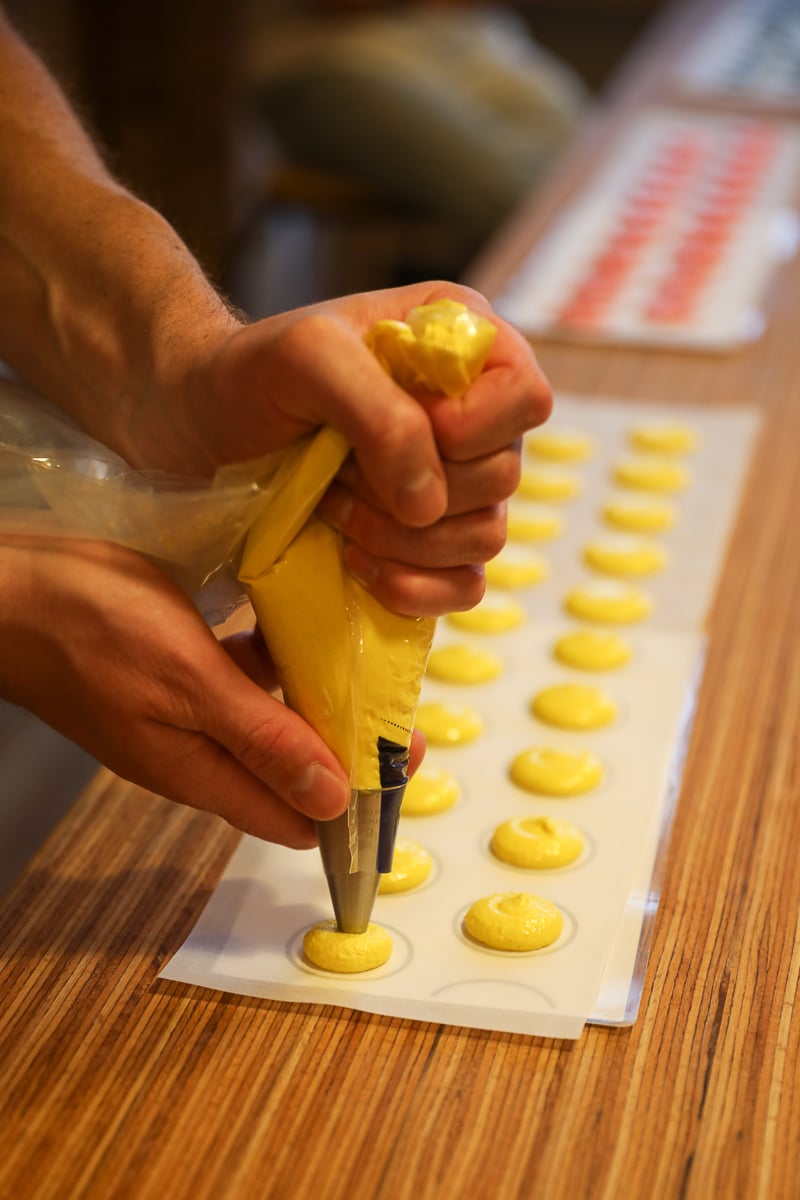 Hands piping yellow macaron batter into even rounds on a guide-lined baking sheet.