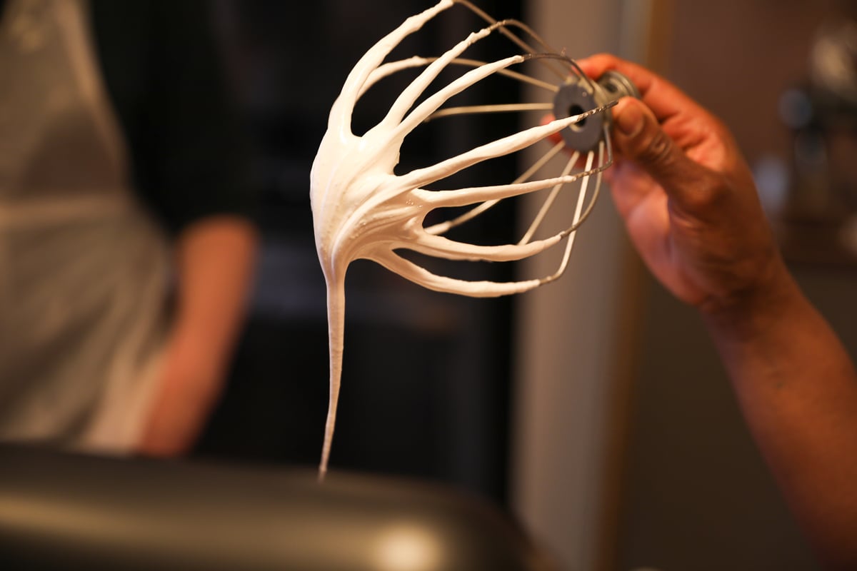 Whisk attachment lifted from a stand mixer trailing glossy Italian meringue in a soft peak.