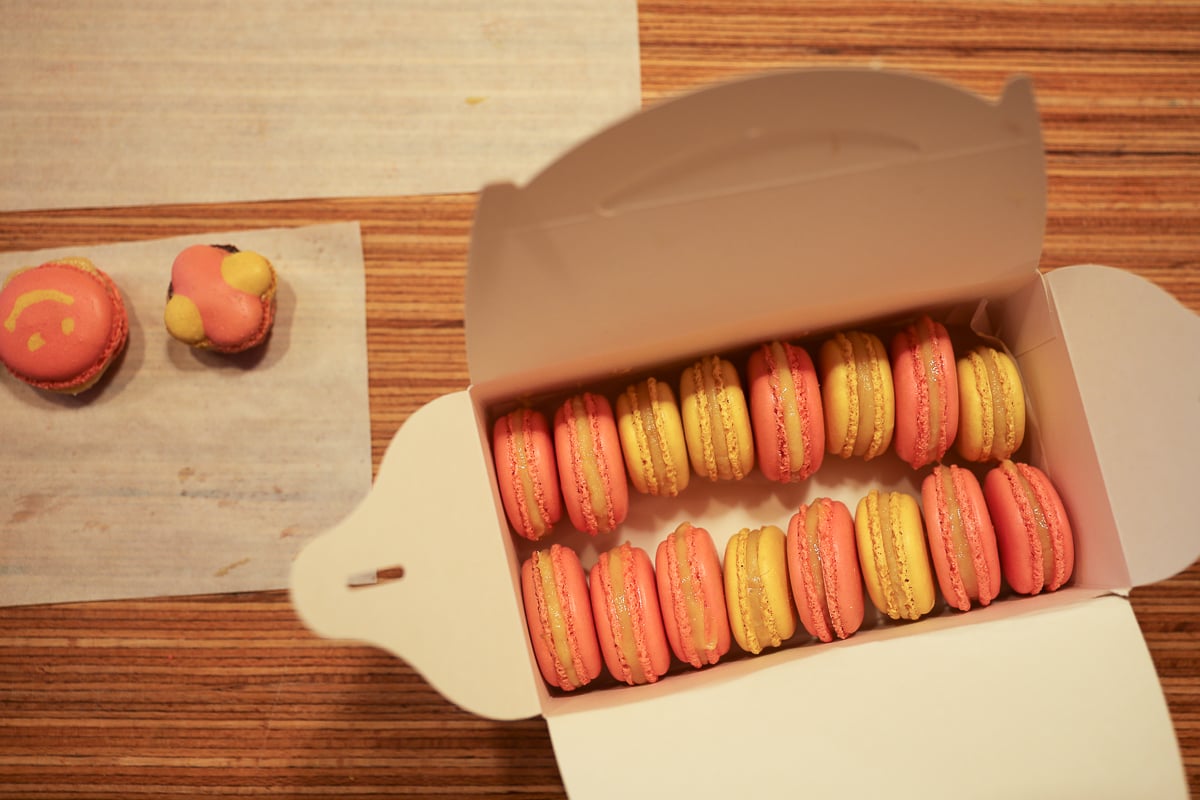 Open box of homemade pink and yellow macarons, with two playfully decorated ones resting beside it.