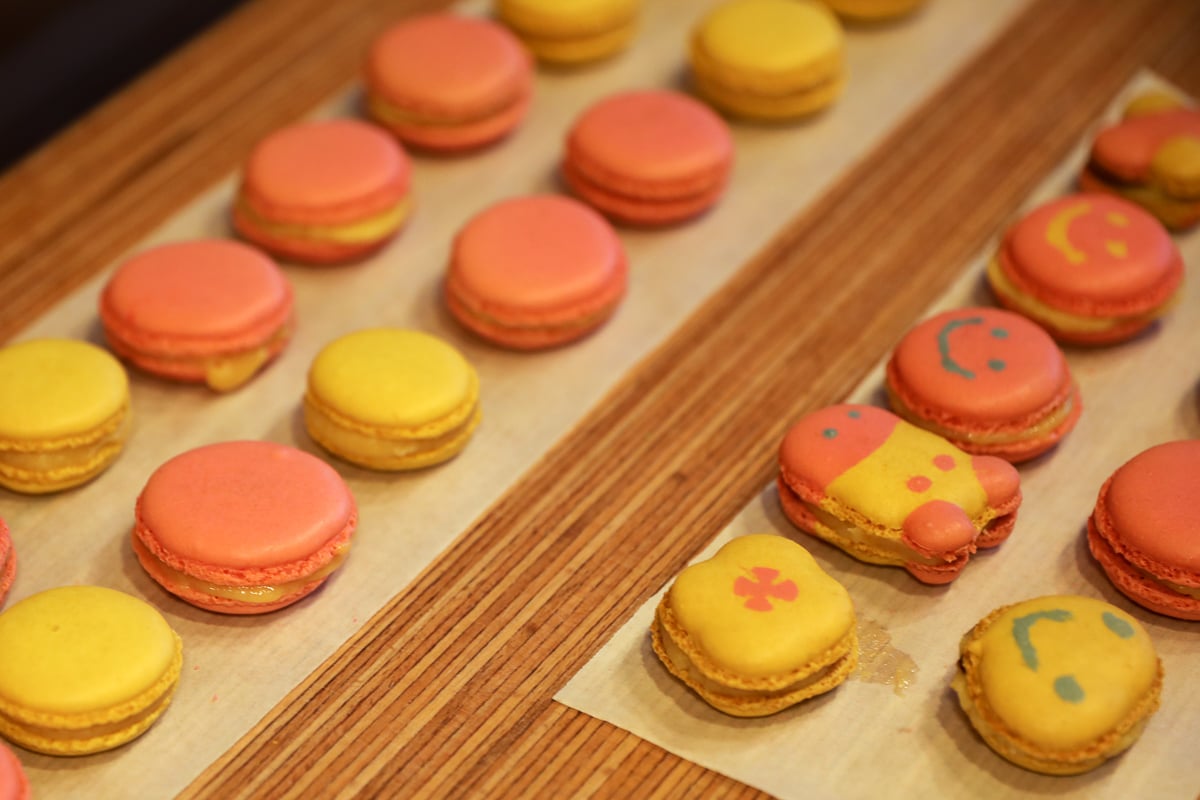 Rows of pink and yellow macarons, including a few decorated with smiley faces and playful designs.
