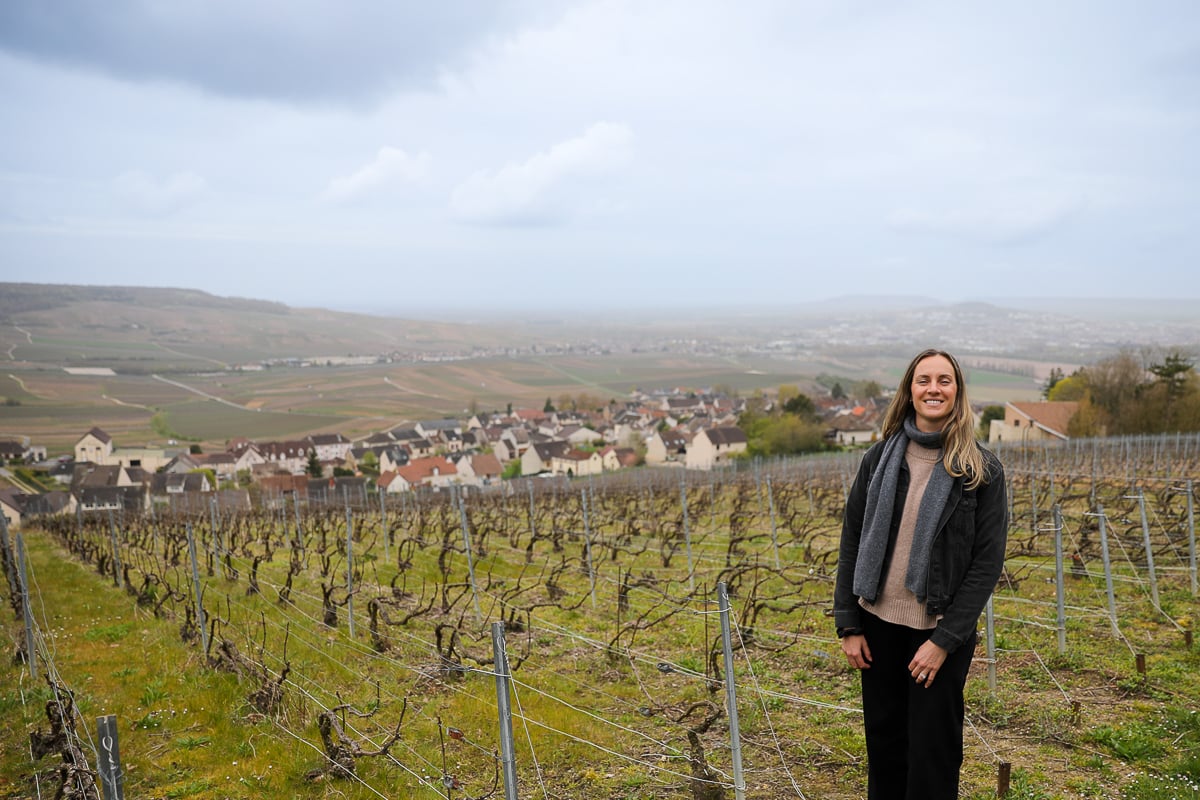 Maddy standing among the vines in a Champagne vineyard with rolling hills beyond